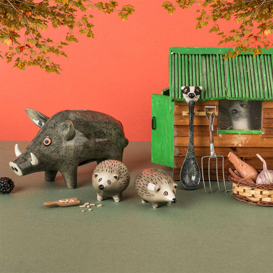 A styled miniature farm scene featuring a small wooden barn with a green roof, a wild boar money box, two hedgehog salt & pepper shakers, badger spoon, a pitchfork, a rake, a broom, a basket with garlic and sweet potatoes, carrots, and a fox egg cup, arranged on a green surface against a coral-orange backdrop with hanging leaves.