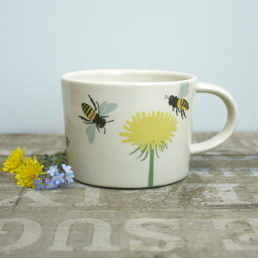 Ceramic mug with bee and dandelion design on a wooden surface with flowers.