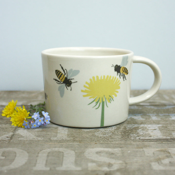 Ceramic mug with bee and dandelion design on a wooden surface with flowers.