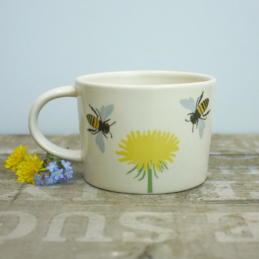 Ceramic mug with bee and dandelion design on a wooden surface with flowers.