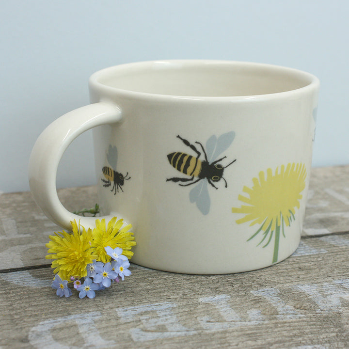 White mug with bee and dandelion design on a wooden surface with flowers.