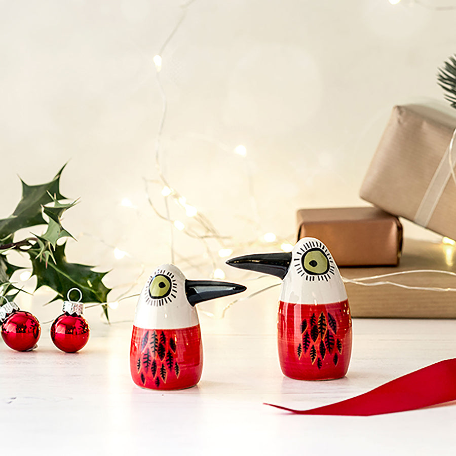 Ceramic  red bird salt and pepper shakers with black beaks and green eyes, displayed on a white surface with holiday decorations including holly, red baubles, wrapped gifts, and warm string lights in the background.