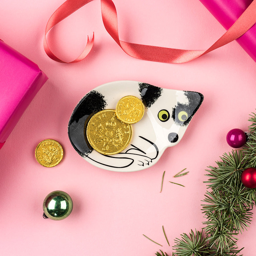A ceramic cat-shaped trinket dish by Hannah Turner holding several silver rings, styled on a pink background with ribbon, pine branches, ornaments and gift boxes.