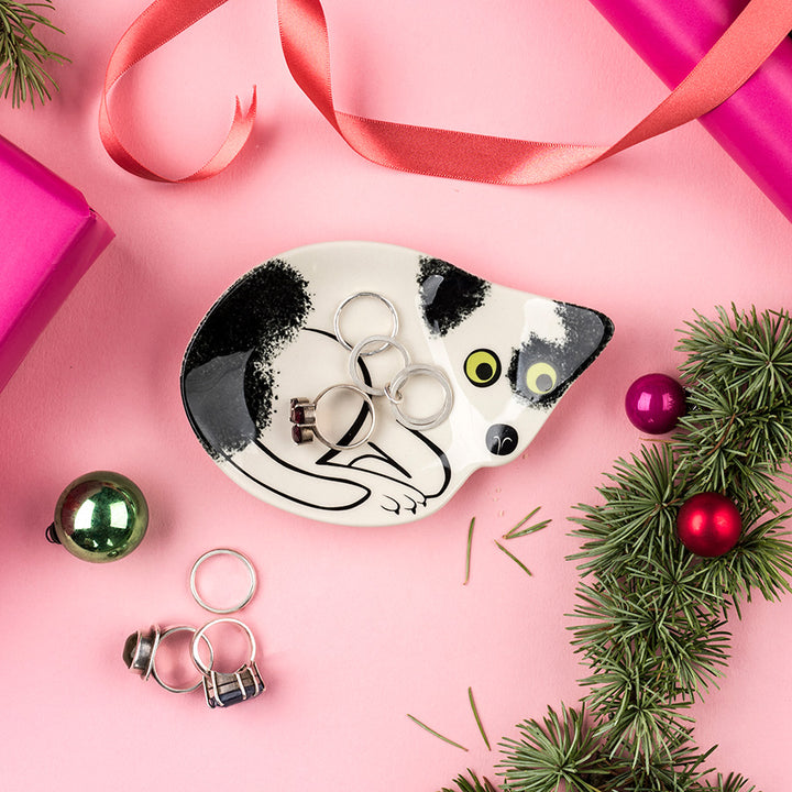 A ceramic cat-shaped trinket dish by Hannah Turner holding several silver rings, styled on a pink background with ribbon, pine branches, ornaments and gift boxes.