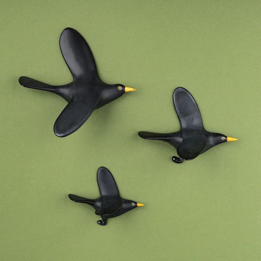 Three black bird wall decorations shaped like flying birds with yellow beaks, arranged on a solid green background.