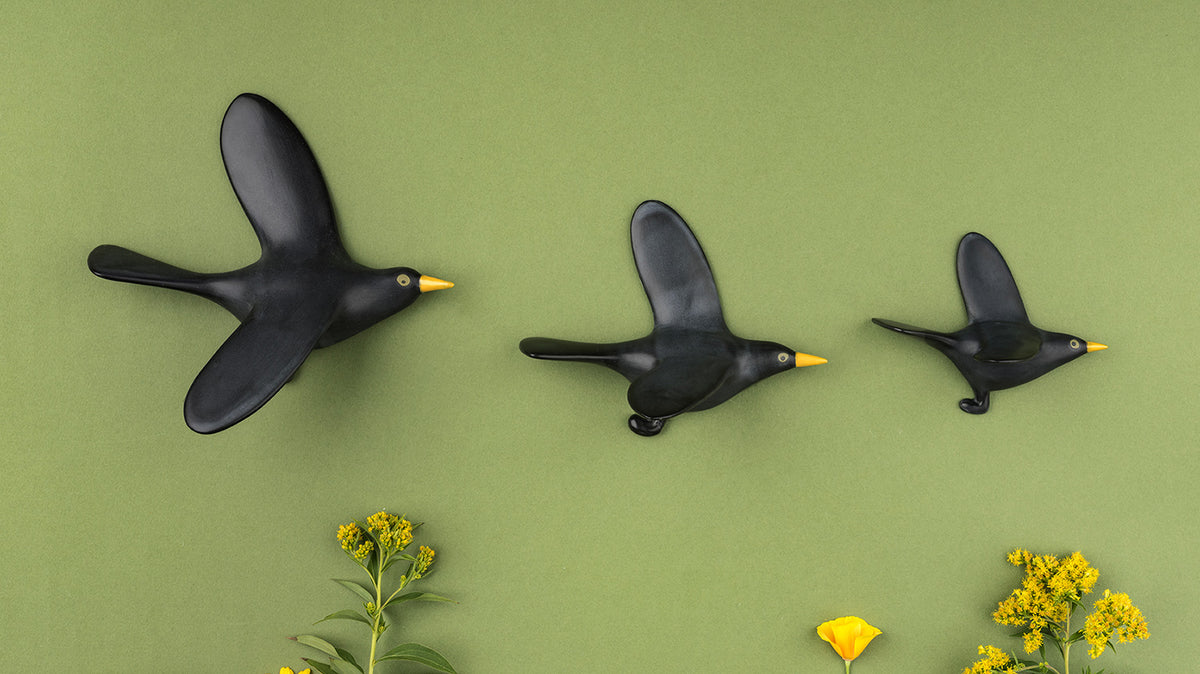 Three ceramic black birds flying with yellow beaks, arranged on a light green background with yellow flowers at the bottom.