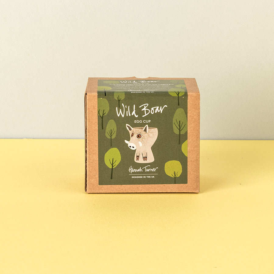 A square kraft paper box for 'Wild Boar Egg Cup' by 'Hannah Turner sits on a yellow surface against a light beige wall. The box features a dark green front label with an illustration of a wild boar and trees, and a white logo.