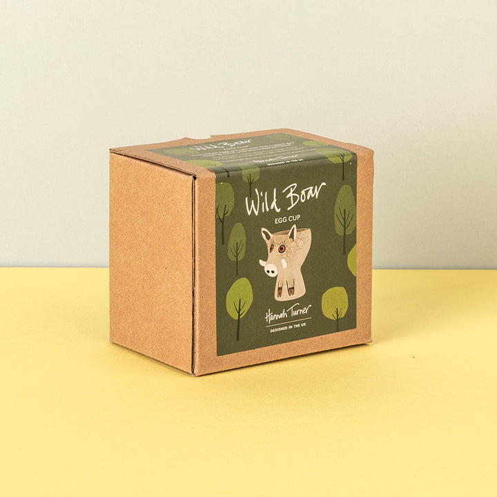 A square kraft paper box with a green illustrated label reading 'Wild Boar Egg Cup' and 'Handmade in the uk' photographed on a pale yellow surface against a light beige wall.