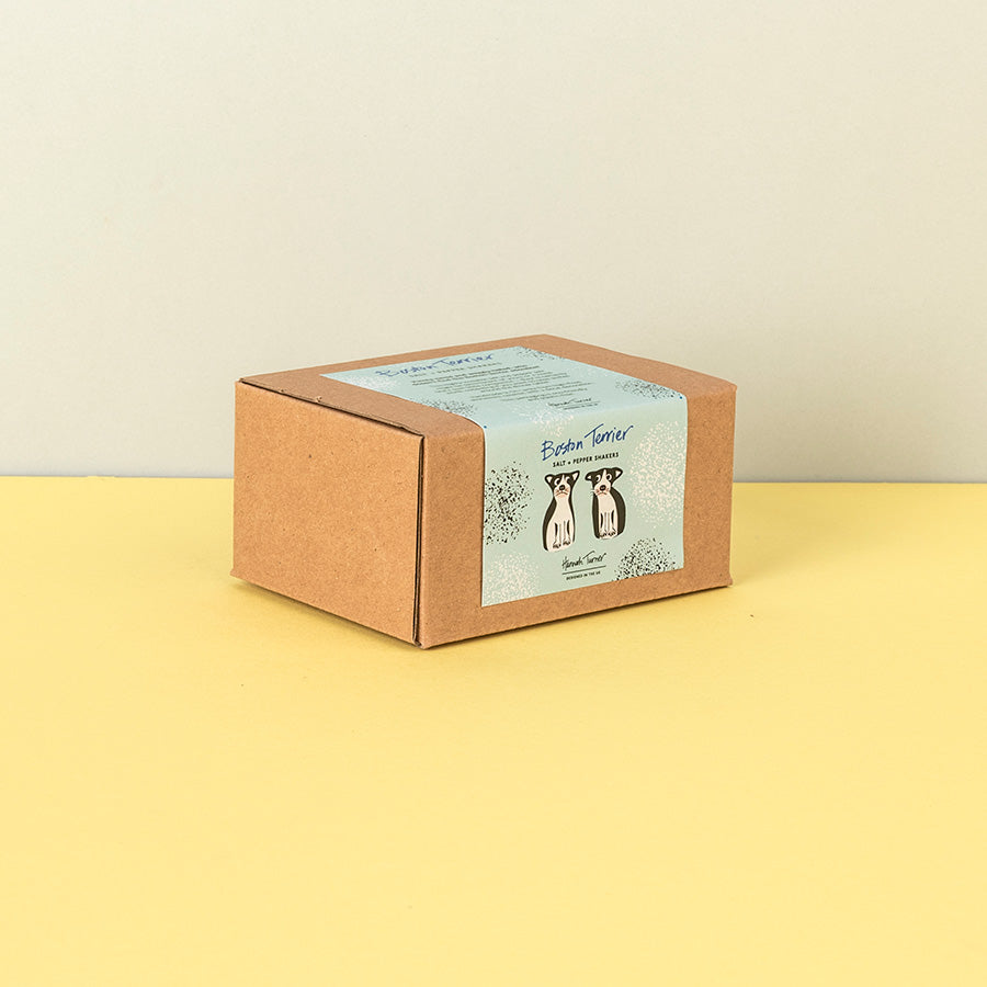 A square brown kraft paper box with a light blue label featuring Boston terrier illustrations and the text 'Salt & Pepper Shakers' and 'Hand Crafted', set on a yellow surface against a beige background.