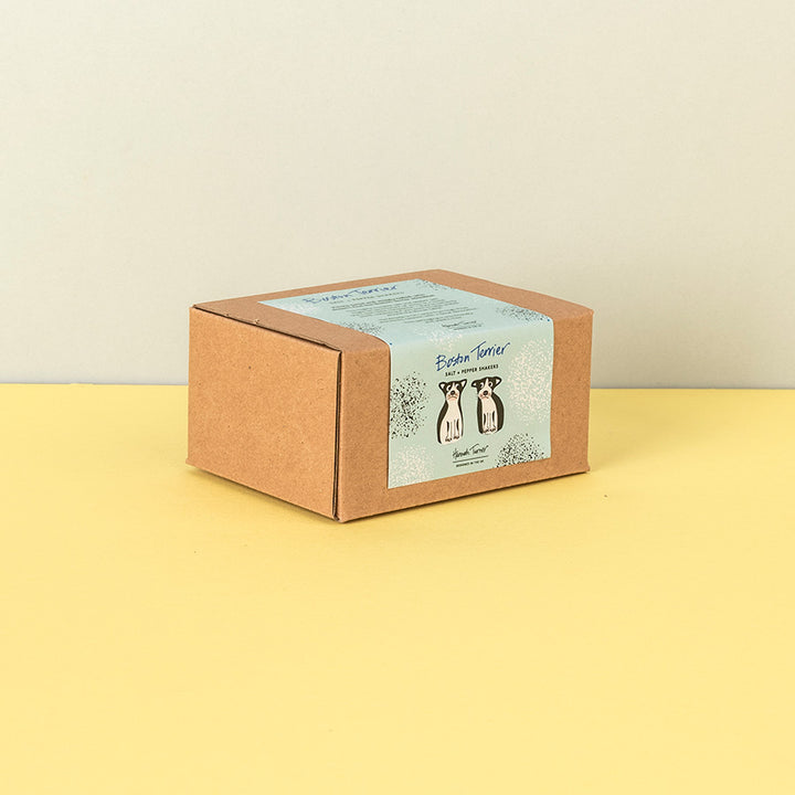 A square brown kraft paper box with a light blue label featuring Boston terrier illustrations and the text 'Salt & Pepper Shakers' and 'Hand Crafted', set on a yellow surface against a beige background.