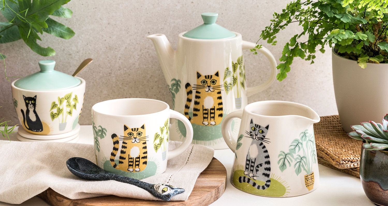 Ceramic tea set with cat designs on a wooden surface with plants in the background