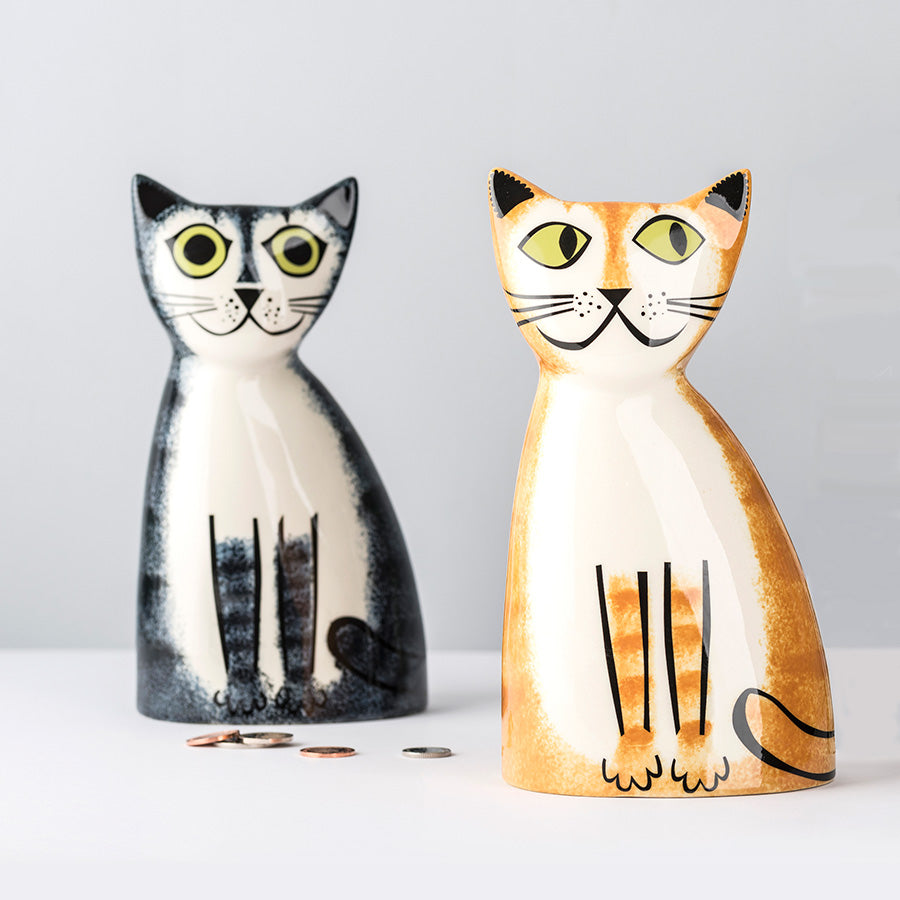 Two ceramic cat-shaped coin jars on a light gray background, one black and white with yellow eyes, and one orange and cream with yellow eyes. A few coins are scattered in front of the black and white cat.