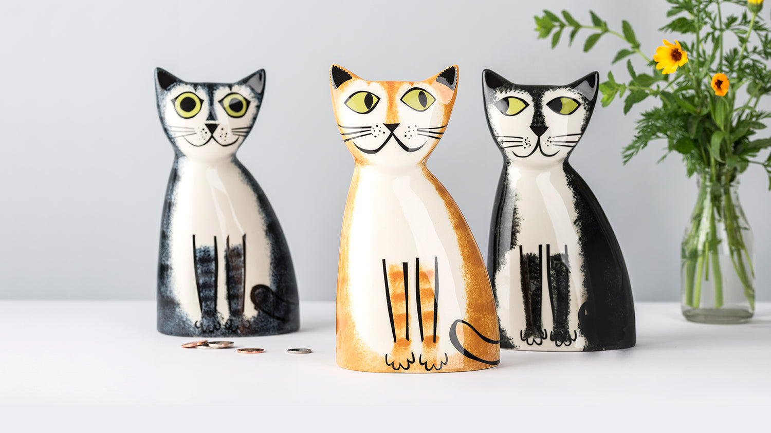 Three cat-shaped money boxes in black, ginger, and grey tabby and black and white  on a light grey background.