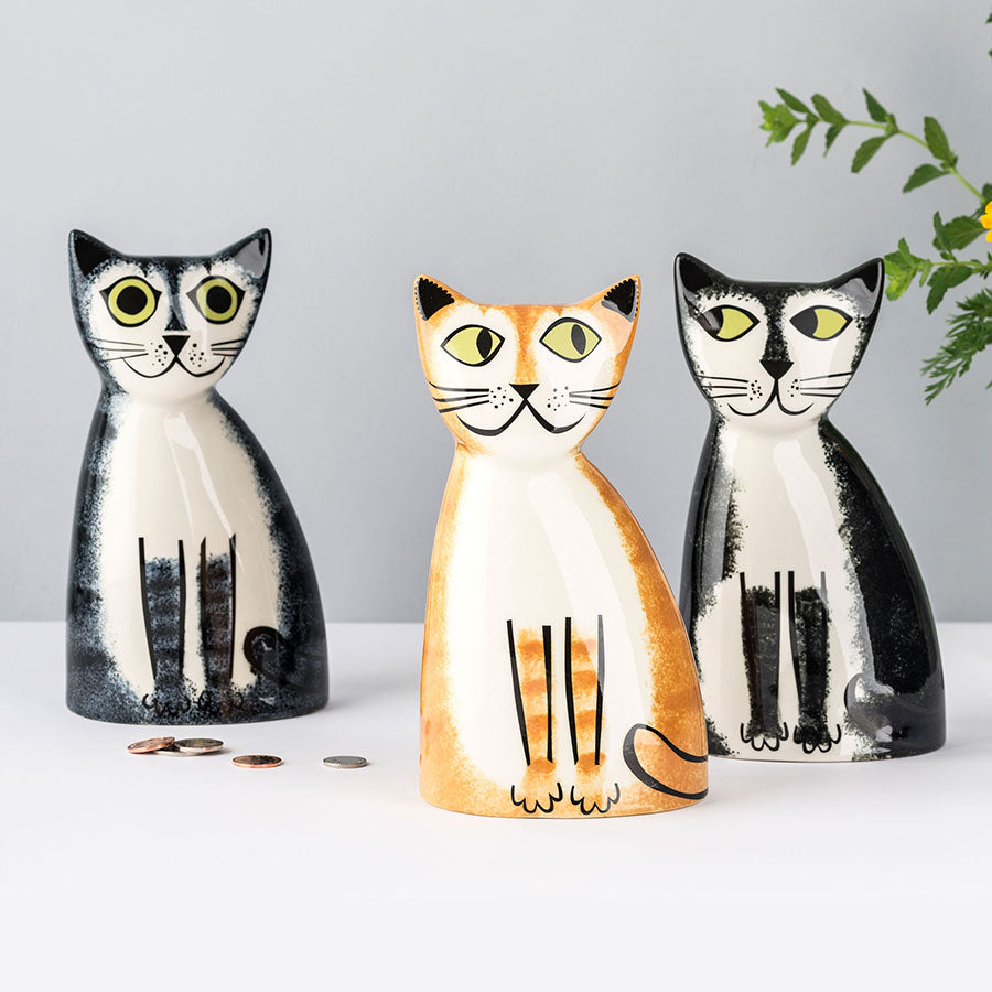 Three cat-shaped money boxes in black, ginger, and grey tabby and black and white  on a light gray background.
