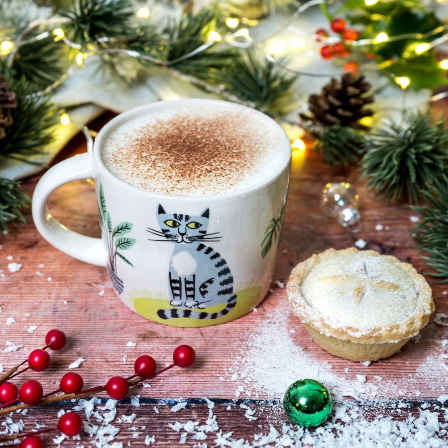 Mug with a cat design, cappuccino, and mince pie on a festive wooden table with Christmas decorations.