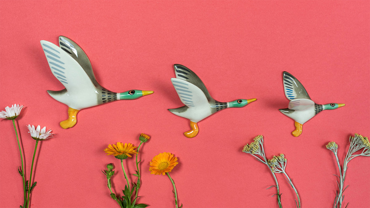 Three decorative ceramic ducks in flight are arranged on a coral pink background with small flowers placed around them. The ducks have white bodies, gray wings, teal heads, and yellow beaks and feet.