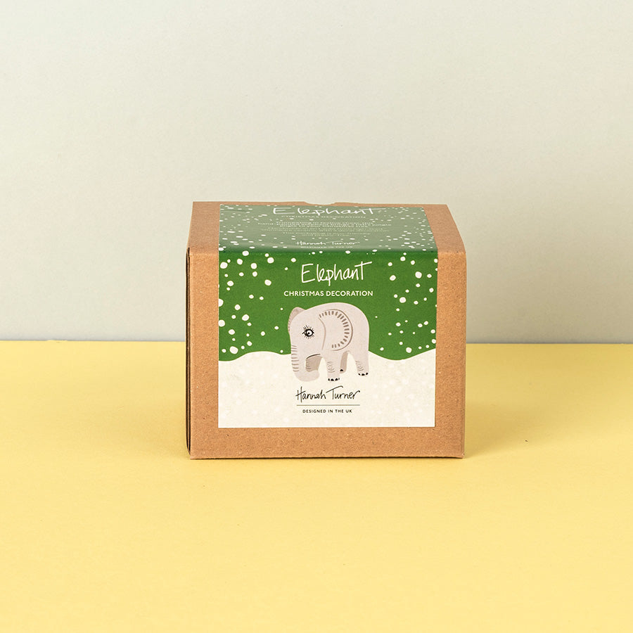 A brown kraft paper box for a 'Elephant Christmas Decoration' by Hannah Turner, featuring a green snowy label with an illustration of a elephant, set against a split beige and yellow background.
