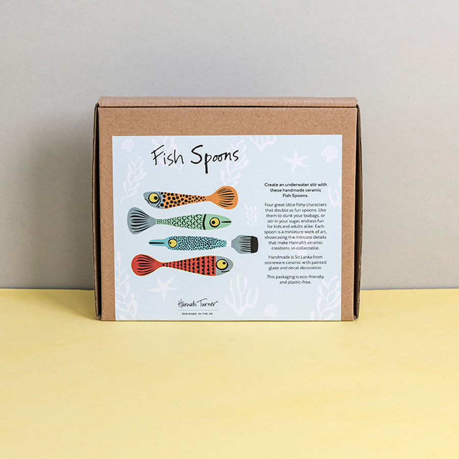 Brown cardboard box of Fish Spoons with illustration on a light grey background with the text 'Hannah Turner designed in the UK'. Packaging shot for Hannah Turner's Ceramic Fish Spoon Set.