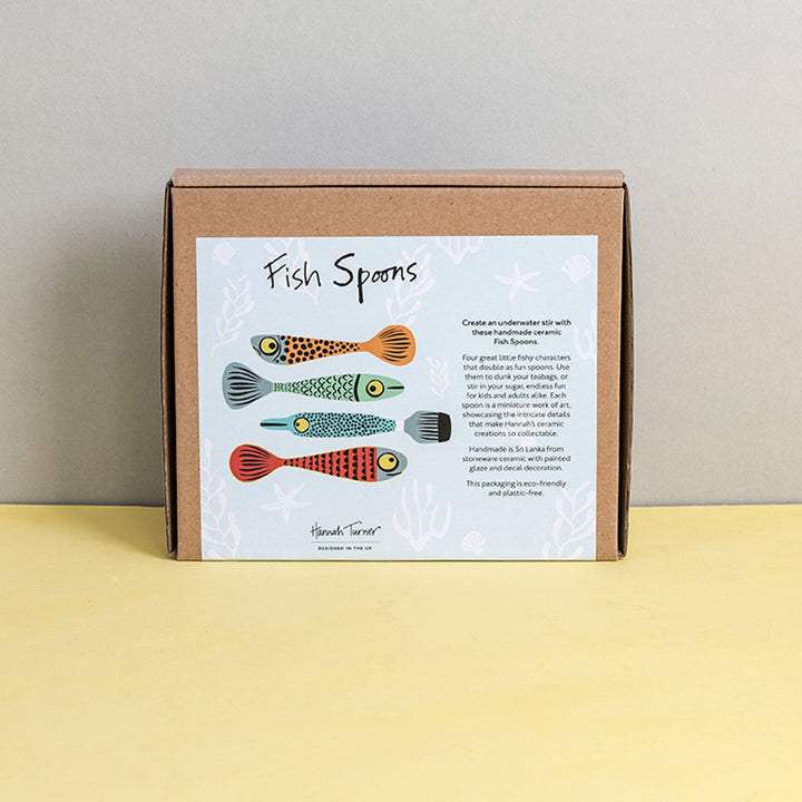 Brown cardboard box of Fish Spoons with illustration on a light grey background with the text 'Hannah Turner designed in the UK'. Packaging shot for Hannah Turner's Ceramic Fish Spoon Set.