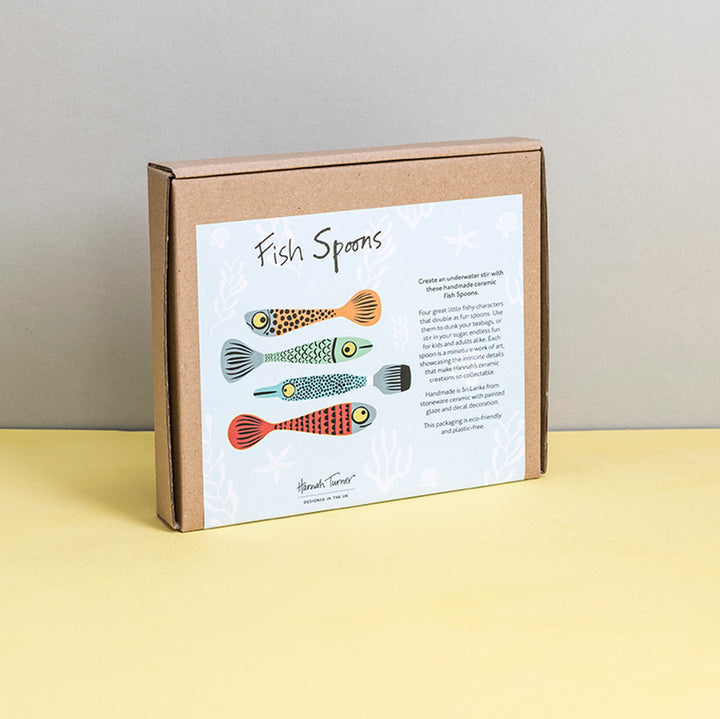 Brown cardboard box of Fish Spoons with illustration on a light grey background with the text 'Hannah Turner designed in the UK'. Packaging shot for Hannah Turner's Ceramic Fish Spoon Set.