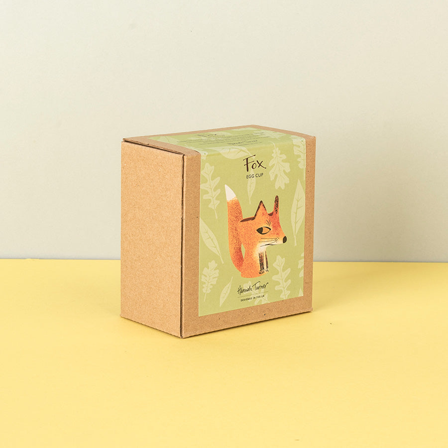 A square kraft paper box with a green illustrated label reading 'Fox Egg Cup' and 'Handmade in the uk' photographed on a pale yellow surface against a light beige wall.