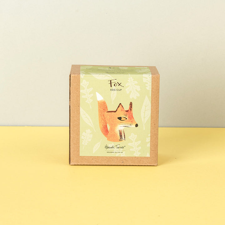 A square kraft paper box with a green illustrated label reading 'Fox Egg Cup' and 'Handmade in the uk' photographed on a pale yellow surface against a light beige wall.