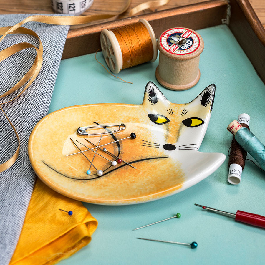 Fox-shaped ceramic trinket dish with pins on a table with spools of thread and fabric.