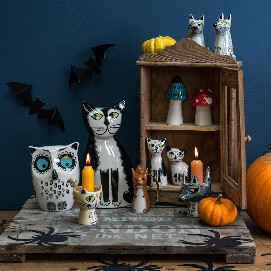 A styled Halloween display featuring ceramics by Hannah Turner in various sizes. A Black & White Cat Money Box, an Owl Money Box, an open wooden cabinet with Toadstool Salt & Pepper Shakers and Black & White Cat Salt & Pepper Shakers inside and Wolf Salt & Pepper Shakers on top. Cat and Wolf Egg Cup, orange candles and pumpkins on a rustic board against a dark blue background with black bat decorations.