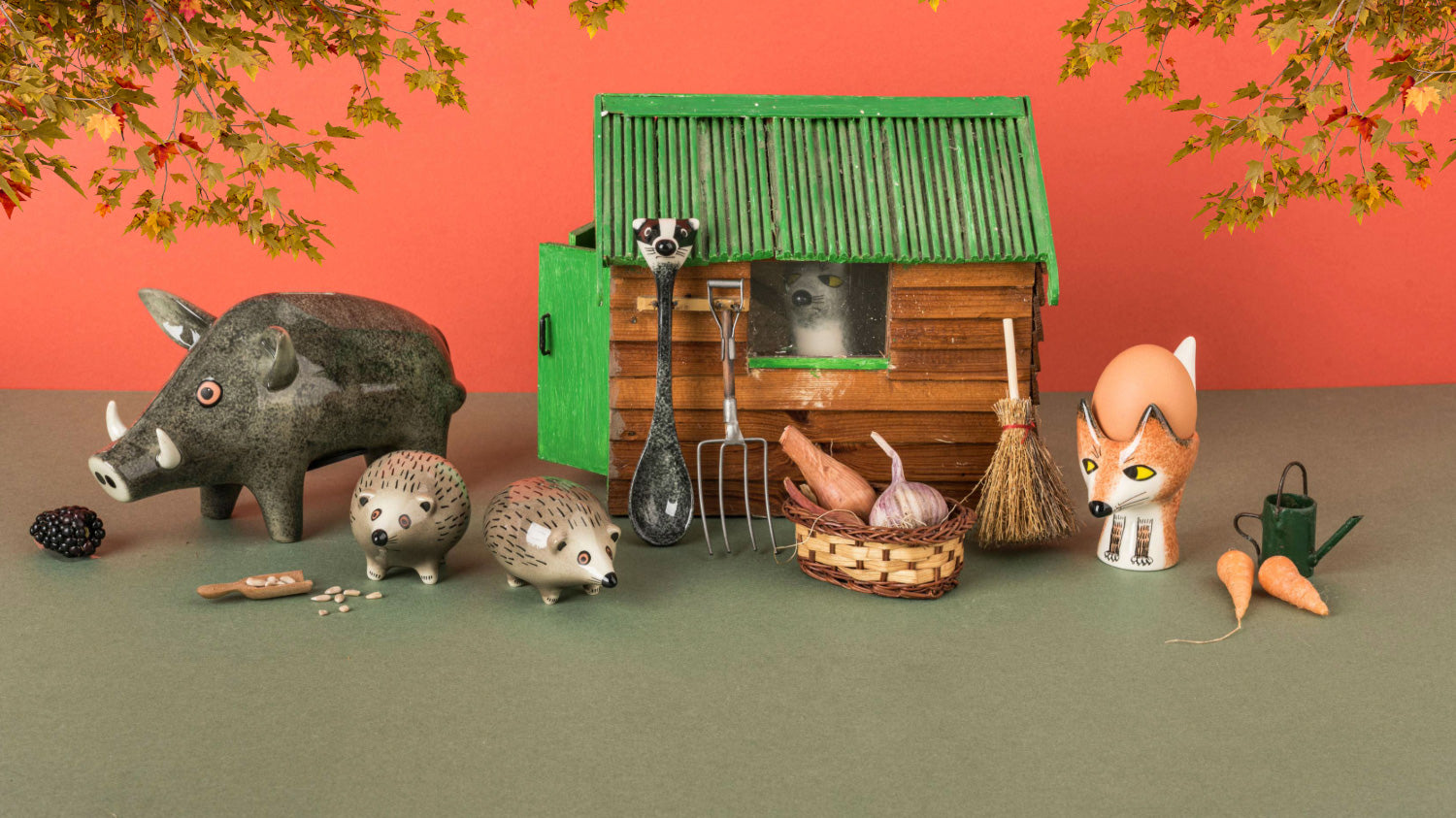 A styled miniature farm scene featuring a small wooden barn with a green roof, a wild boar money box, two hedgehog salt & pepper shakers, badger spoon, a pitchfork, a rake, a broom, a basket with garlic and sweet potatoes, carrots, and a fox egg cup, arranged on a green surface against a coral-orange backdrop with hanging leaves.