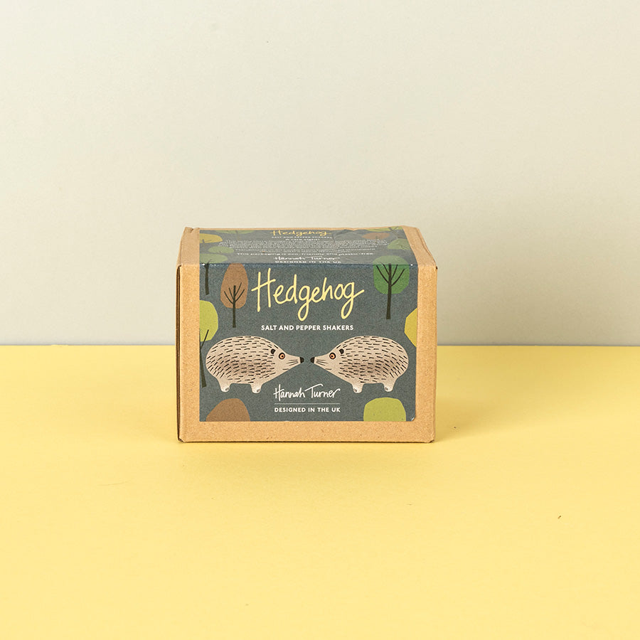 A square kraft paper box of Hedgehog Salt and Pepper Shakers by Hannah Turner, featuring an illustration of two hedgehogs on a muted blue-gray label, set against a split beige and pale yellow background.