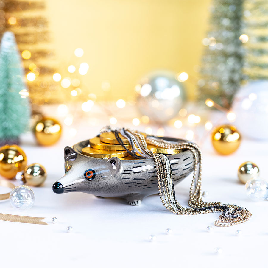 A hedgehog-shaped trinket dish filled with gold coins and draped with layered gold and silver necklaces, styled on a white surface with festive holiday lights and ornaments in the background.