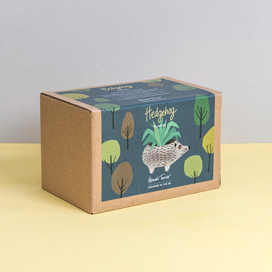 Brown kraft gift box with hedgehog design and trees on a dark grey background printed with text 'Hannah Turner designed in the UK'. Packaging for Hannah Turner's Ceramic Hedgehog Planter.