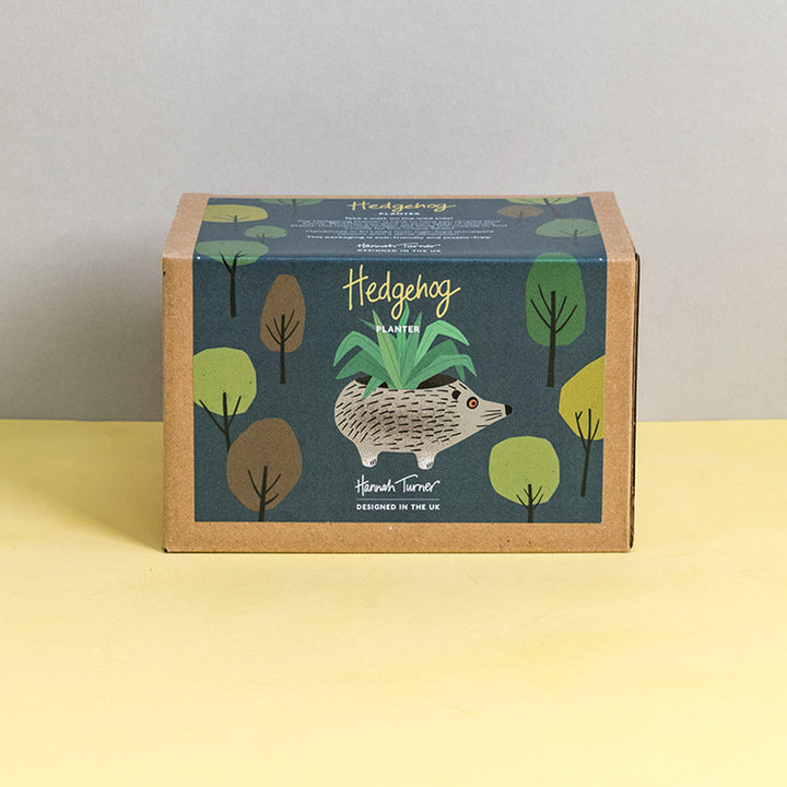 Brown kraft gift box with hedgehog design and trees on a dark grey background printed with text 'Hannah Turner designed in the UK'. Packaging for Hannah Turner's Ceramic Hedgehog Planter.