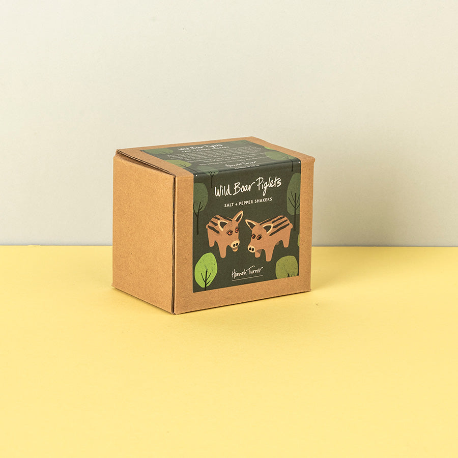 A square kraft paper box of Handmade Soap's 'Wild Boar Piglets' salt and pepper shakers, featuring an illustration of two brown bears on a dark green label, set against a split yellow and beige background.