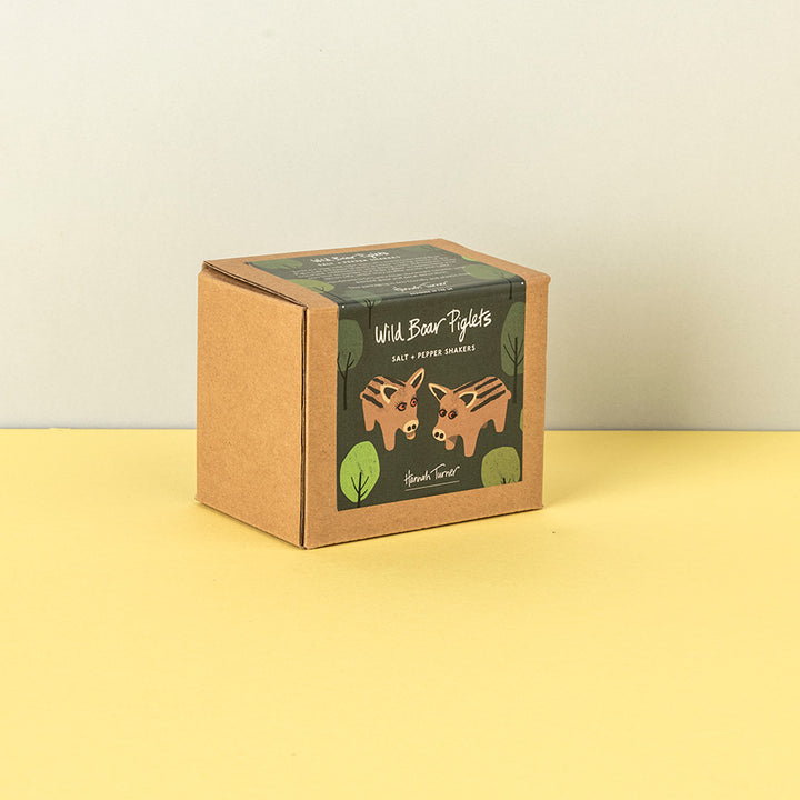 A square kraft paper box of Handmade Soap's 'Wild Boar Piglets' salt and pepper shakers, featuring an illustration of two brown bears on a dark green label, set against a split yellow and beige background.