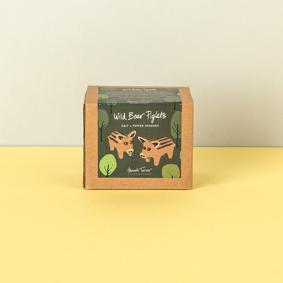 A square kraft paper box of Handmade Soap's 'Wild Boar Piglets' salt and pepper shakers, featuring an illustration of two brown bears on a dark green label, set against a split yellow and beige background.