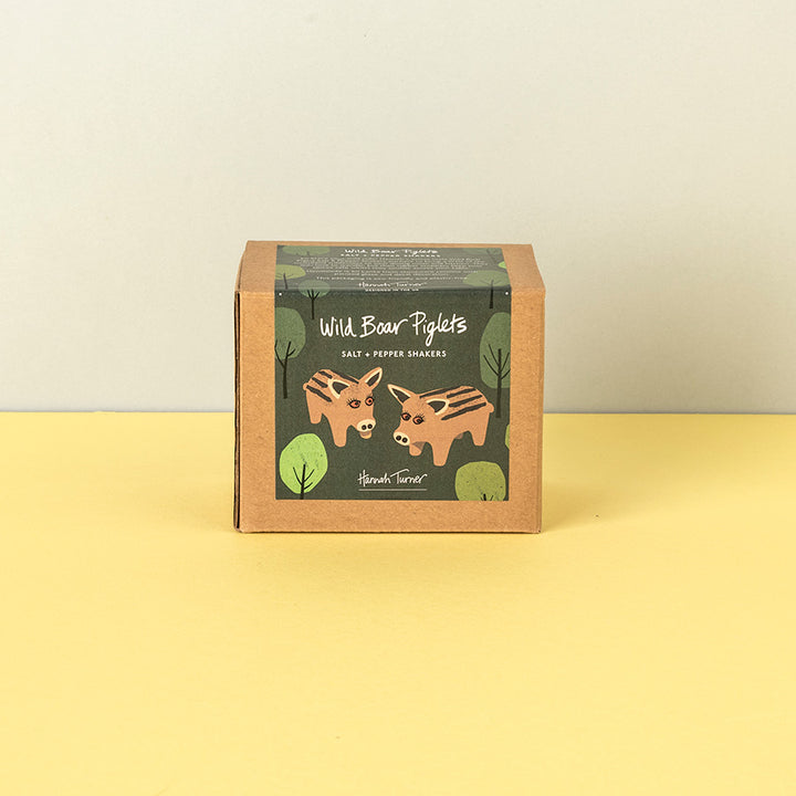 A square kraft paper box of Handmade Soap's 'Wild Boar Piglets' salt and pepper shakers, featuring an illustration of two brown bears on a dark green label, set against a split yellow and beige background.