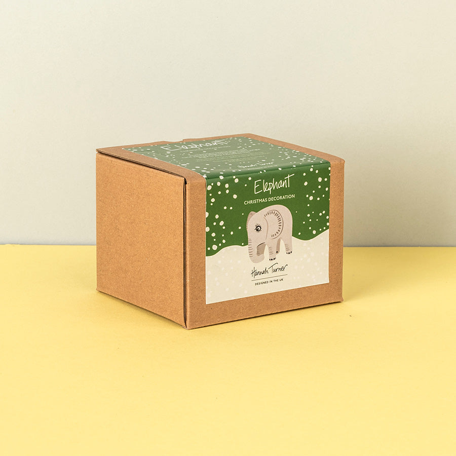 A brown kraft paper box for a 'Elephant Christmas Decoration' by Hannah Turner, featuring a green snowy label with an illustration of a elephant, set against a split beige and yellow background.