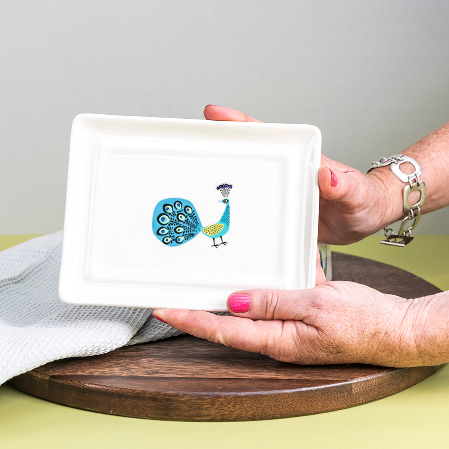 Person the bottom of the butter dish with a peacock design on a wooden surface.