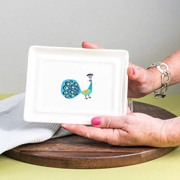 Person the bottom of the butter dish with a peacock design on a wooden surface.