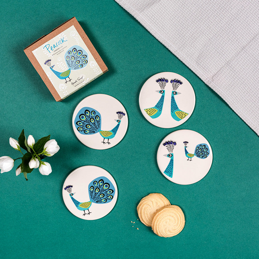 Set of peacock coasters on a green surface with a box and flowers.