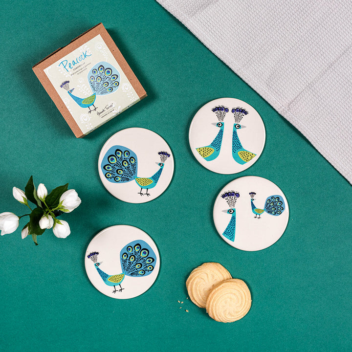 Set of peacock coasters on a green surface with a box and flowers.