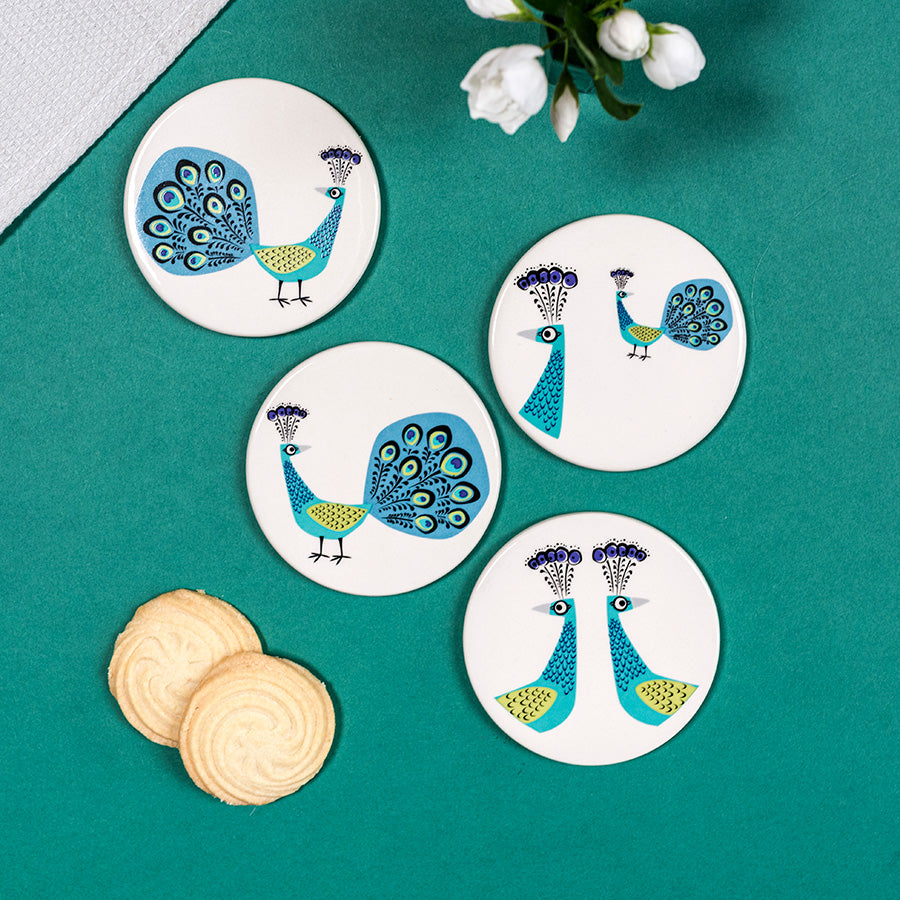 Four peacock-themed coasters on a teal surface with cookies and flowers.