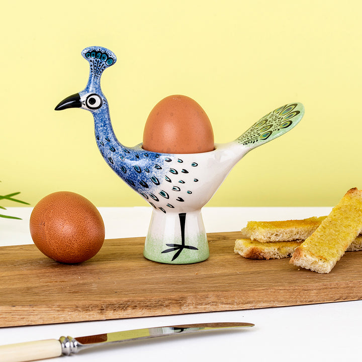 Decorative ceramic peacock-shaped egg cup with an egg on a wooden board.