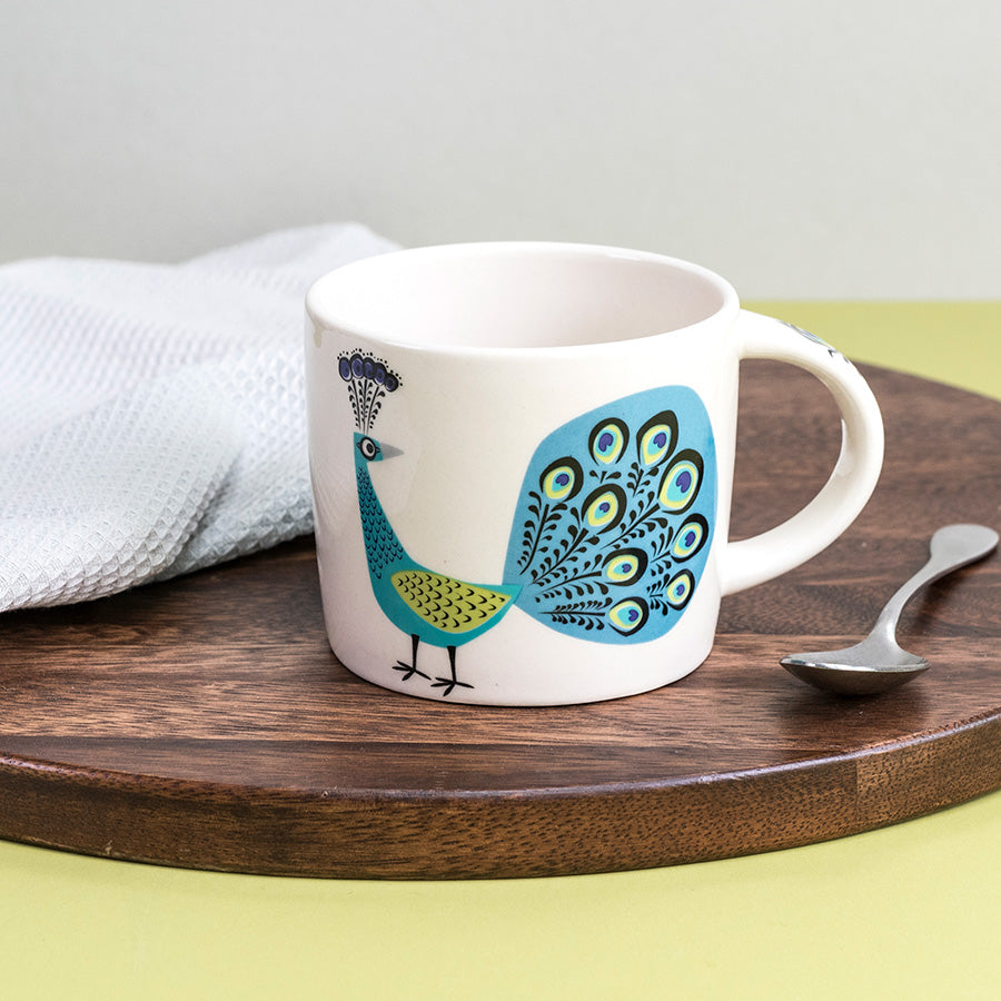 White mug with peacock design on a wooden tray with a spoon and white towel.