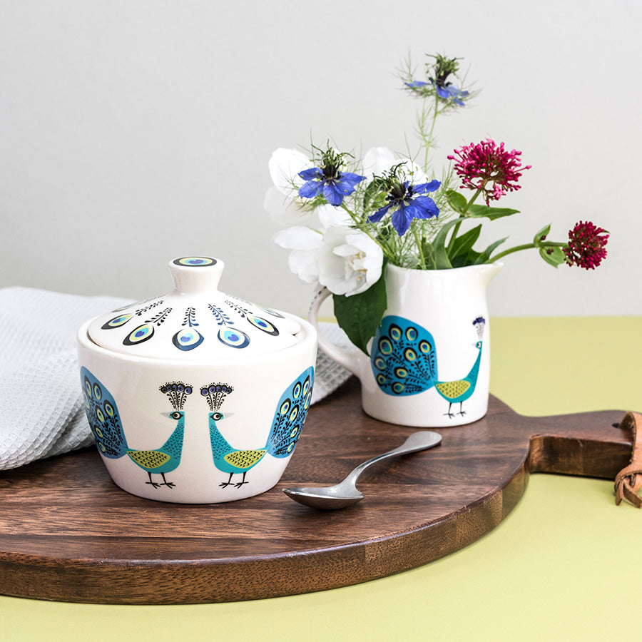 Peacock Sugar Bowl sits on a dark wooden chopping board, with a tea spoon, behind is a milk jugs full of flowers