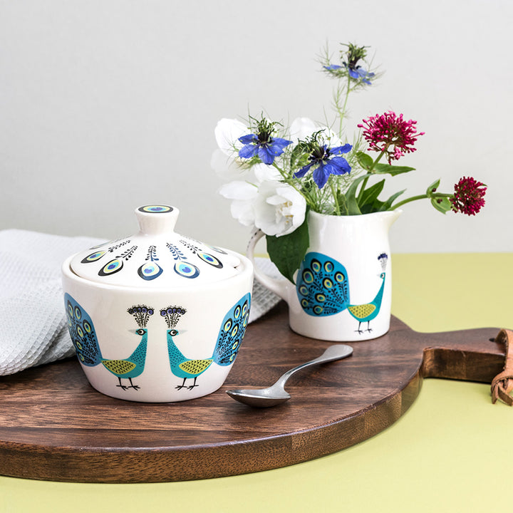 Peacock Sugar Bowl sits on a dark wooden chopping board, with a tea spoon, behind is a milk jugs full of flowers