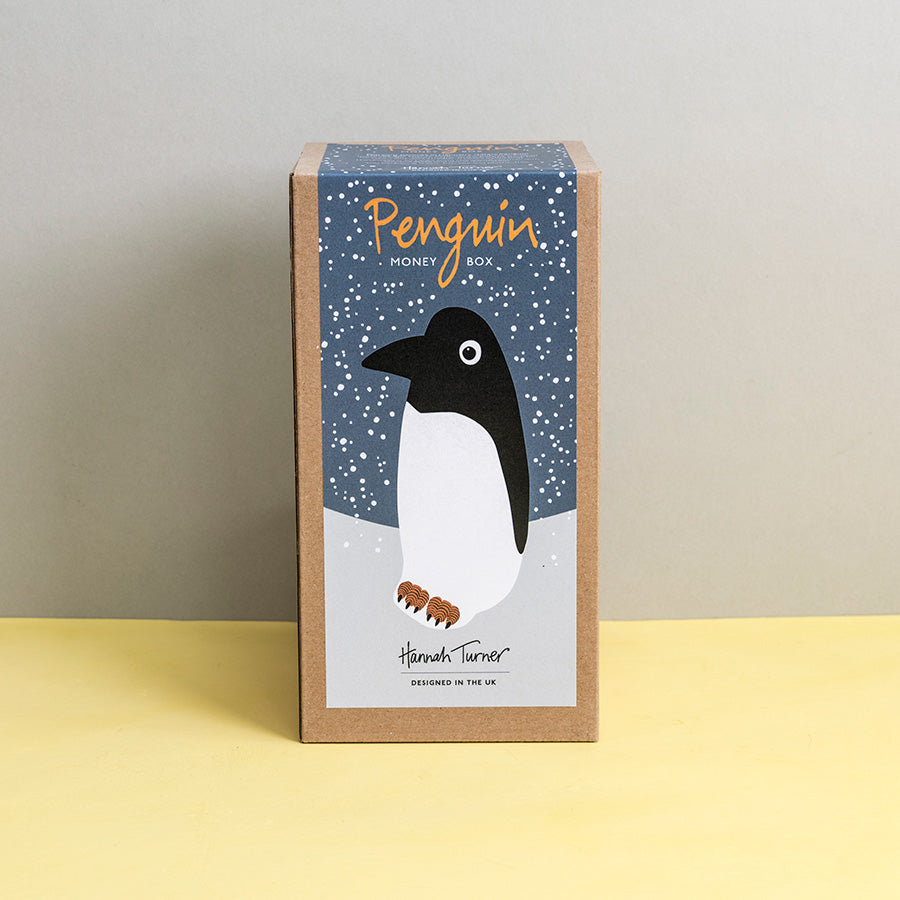 Brown kraft gift box with penguin design on a grey snowy background printed with text 'Hannah Turner designed in the UK'. Packaging for Hannah Turner's Ceramic Penguin Money Box.