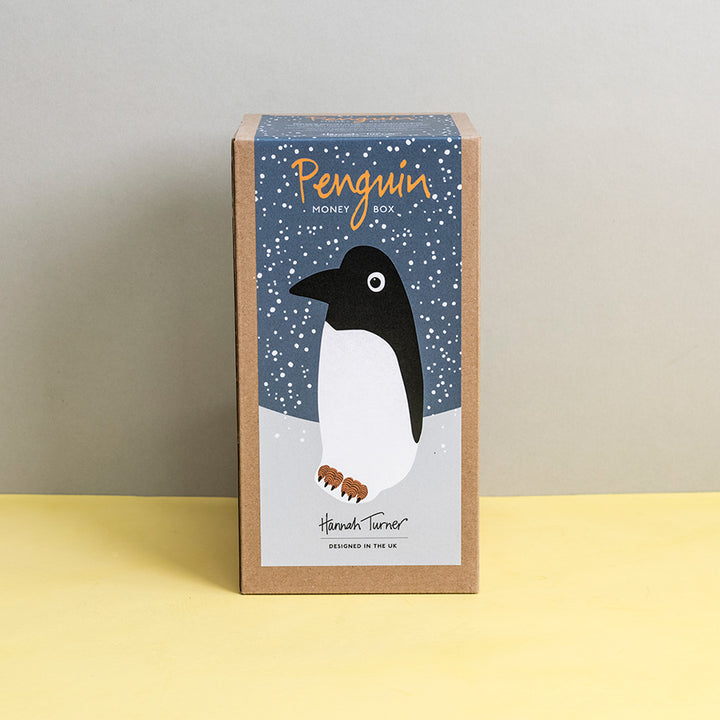 Brown kraft gift box with penguin design on a grey snowy background printed with text 'Hannah Turner designed in the UK'. Packaging for Hannah Turner's Ceramic Penguin Money Box.