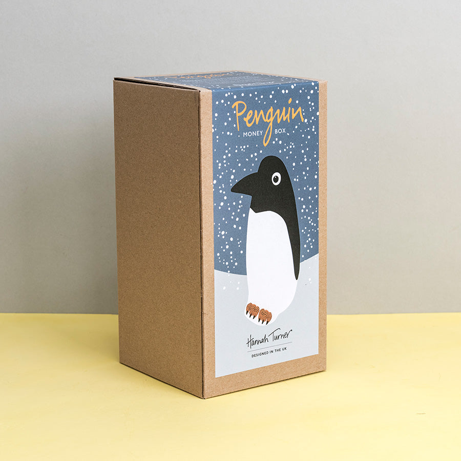 Brown kraft gift box with penguin design on a grey snowy background printed with text 'Hannah Turner designed in the UK'. Packaging for Hannah Turner's Ceramic Penguin Money Box.